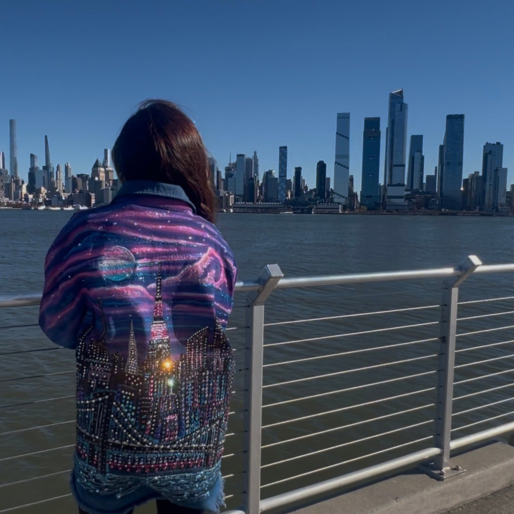 Person wearing a colorful Loria NYC Art jacket looking at a city skyline from a waterfront.