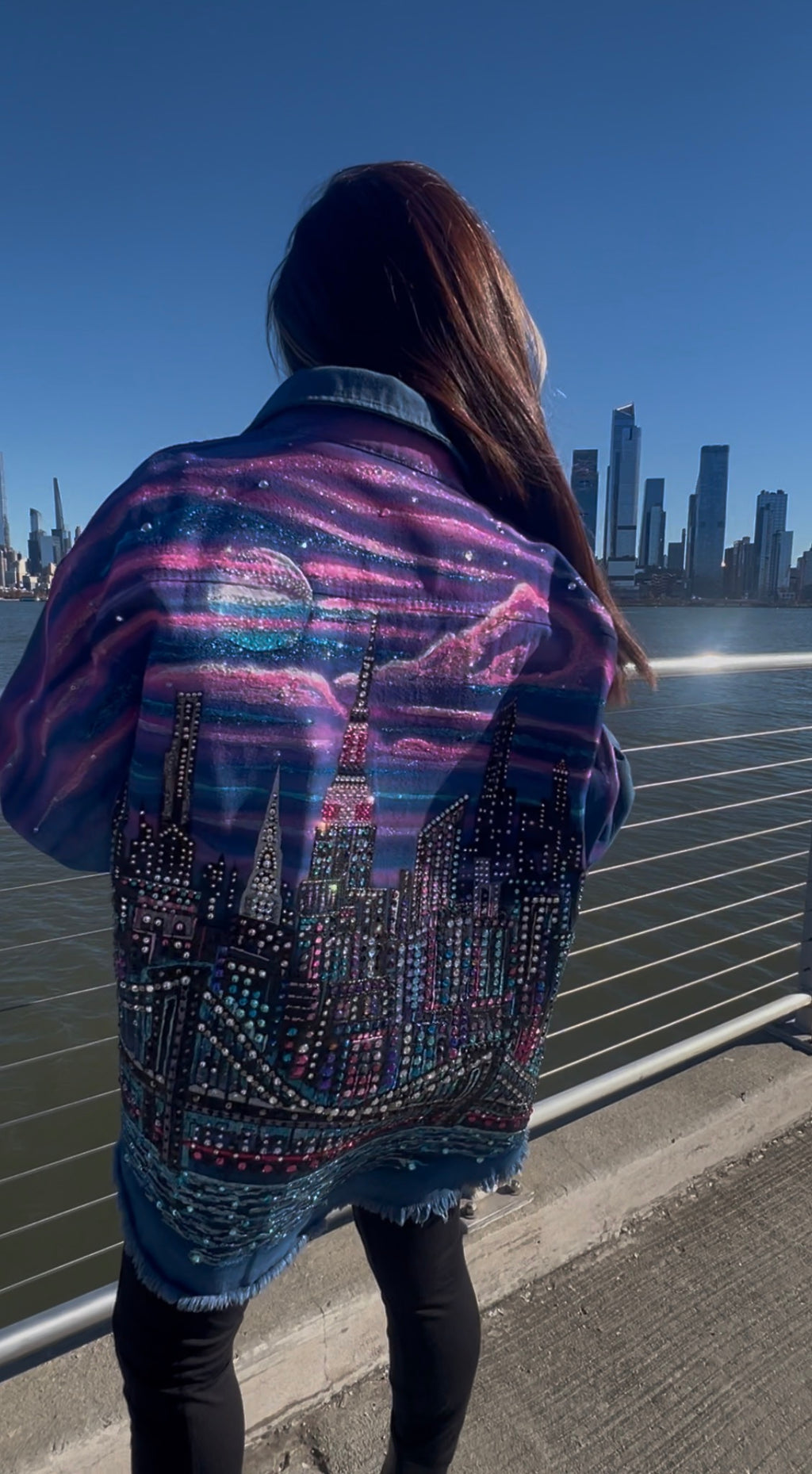 Person wearing a jacket with a Loria NYC Art cityscape design by a waterfront with buildings in the background