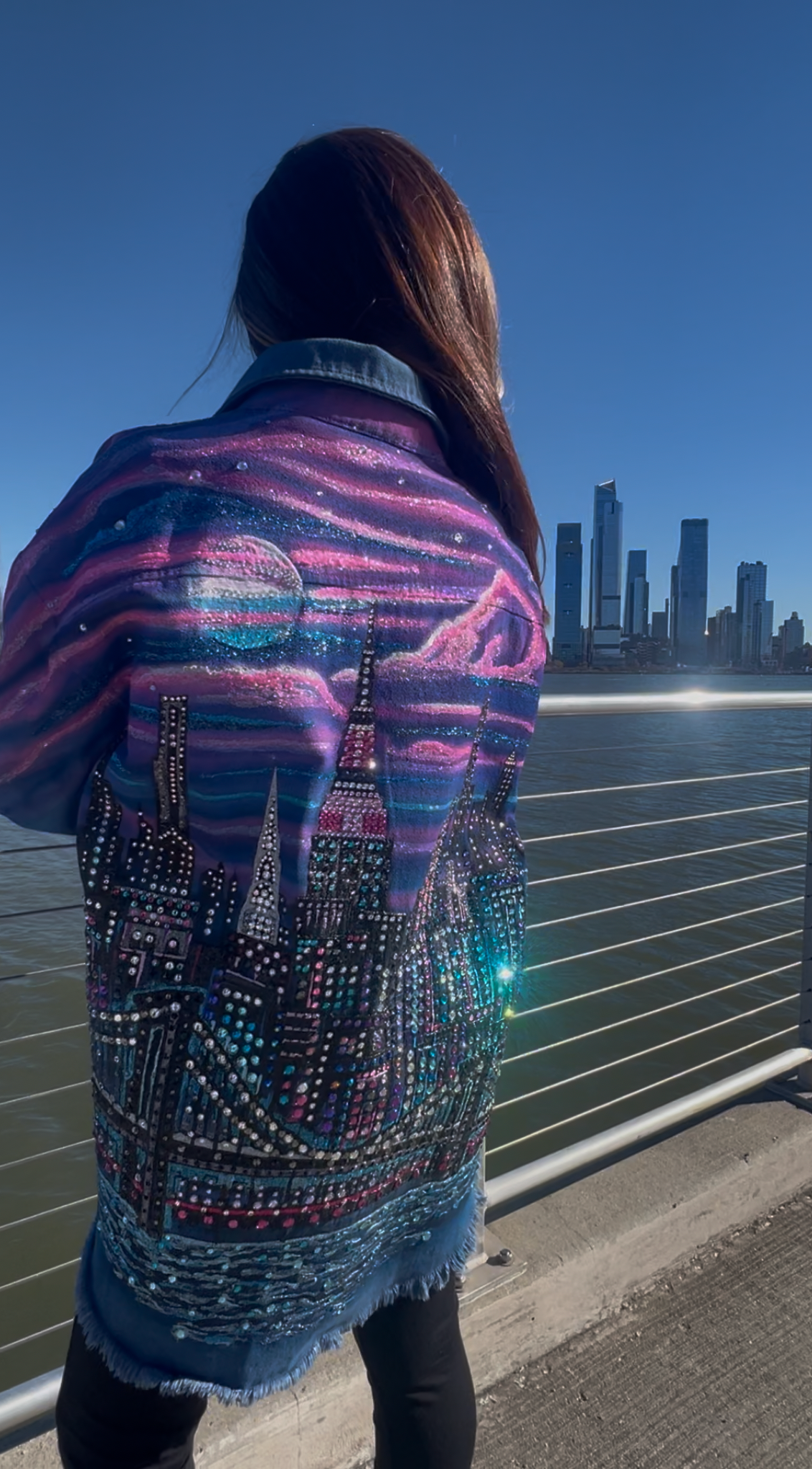 Person wearing a colorful jacket with cityscape Loria NYC Art design, standing on a bridge at night.