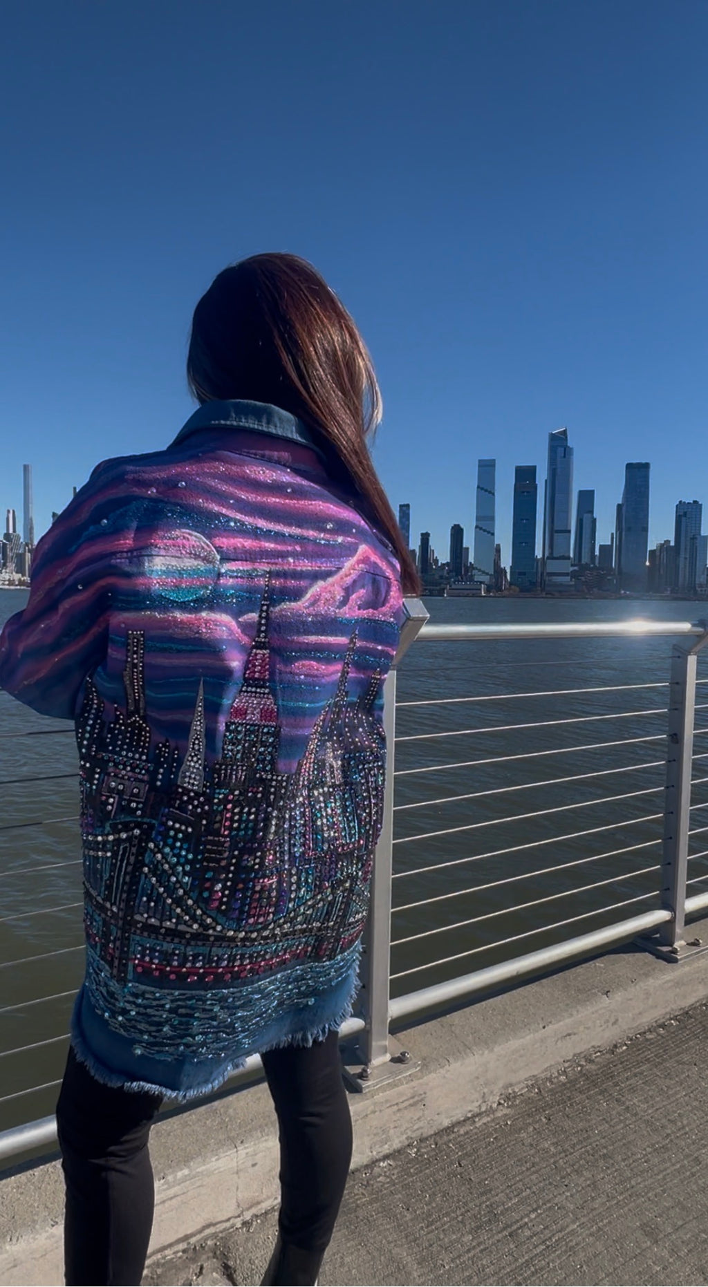 Person wearing a colorful jacket with cityscape Loria NYC Art design, standing by a waterfront with buildings in the background.