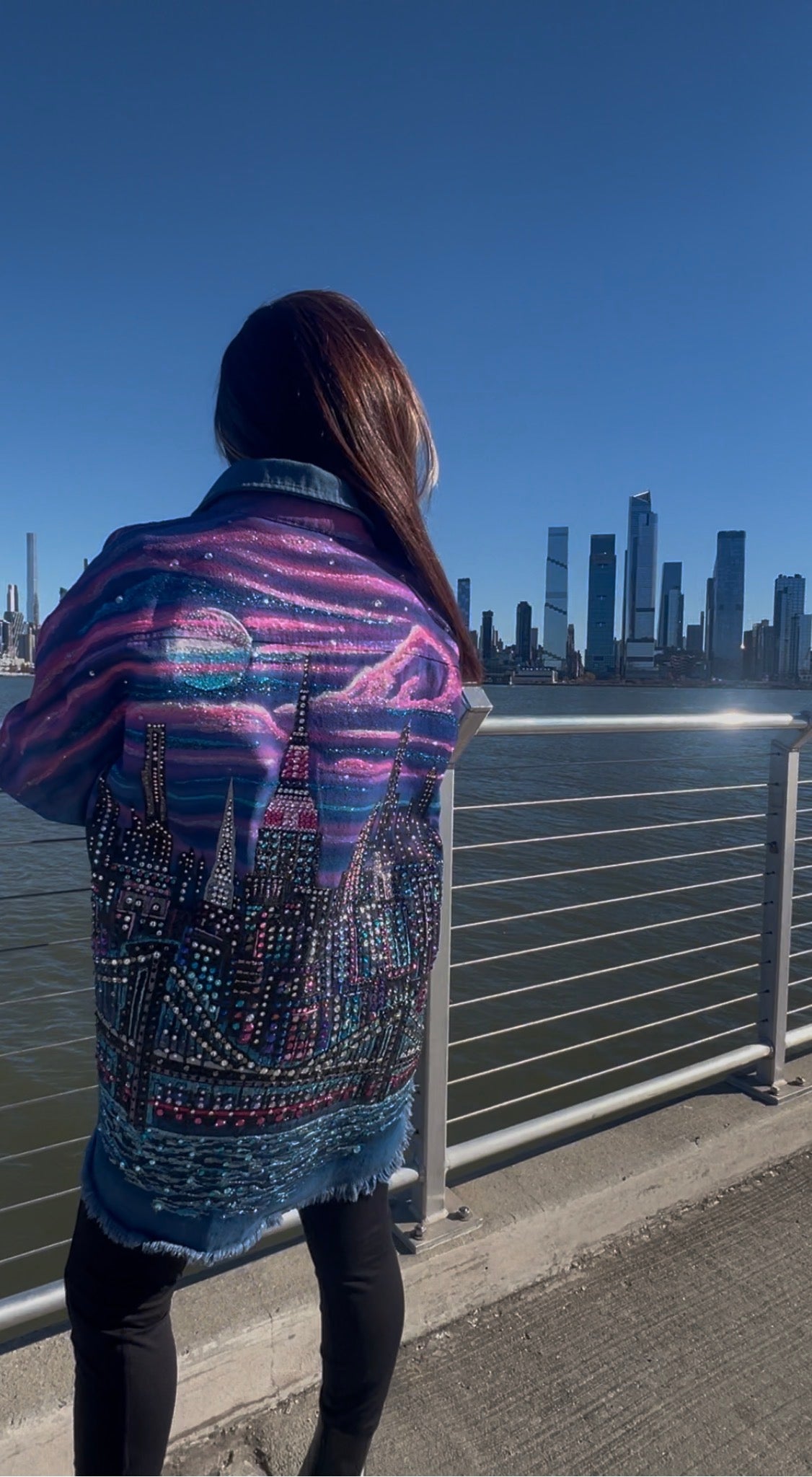 Person wearing a colorful jacket with cityscape Loria NYC Art design, standing by a waterfront with buildings in the background.