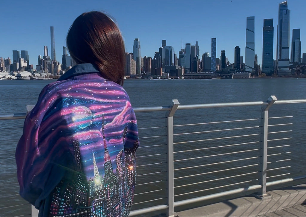 Person wearing a colorful Loria NYC Art jacket looking at a city skyline across water