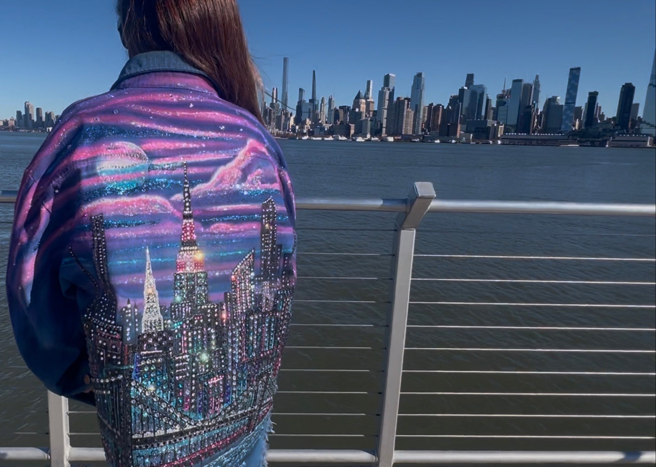 Person wearing a jacket with a Loria NYC Art cityscape design by a waterfront with a city skyline in the background.