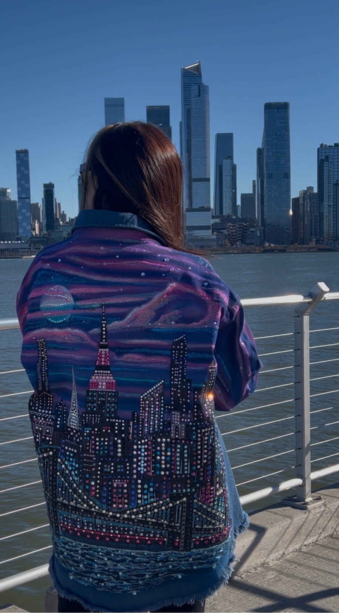 Person wearing a denim jacket with cityscape Loria NYC Art design, standing by a waterfront with skyscrapers in the background.
