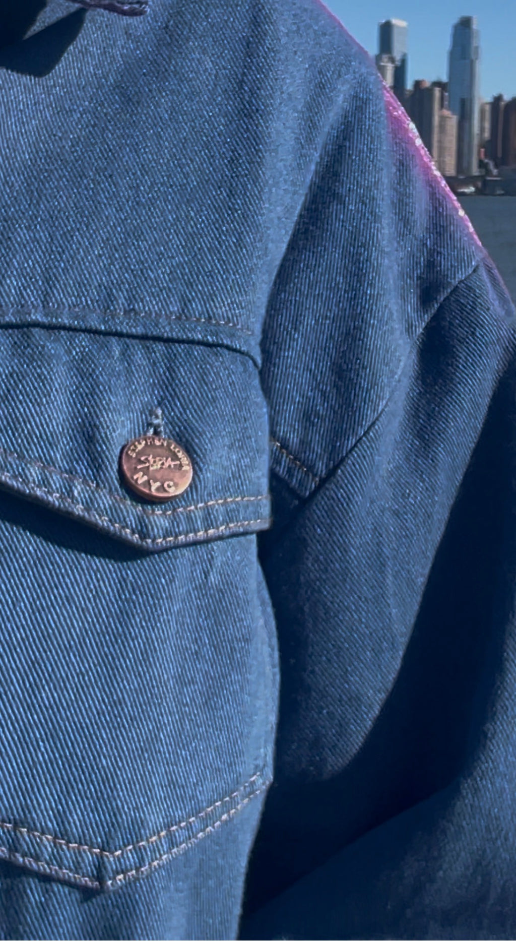 Close-up of a blue denim jacket with a Loria NYC Art button, city skyline in the background