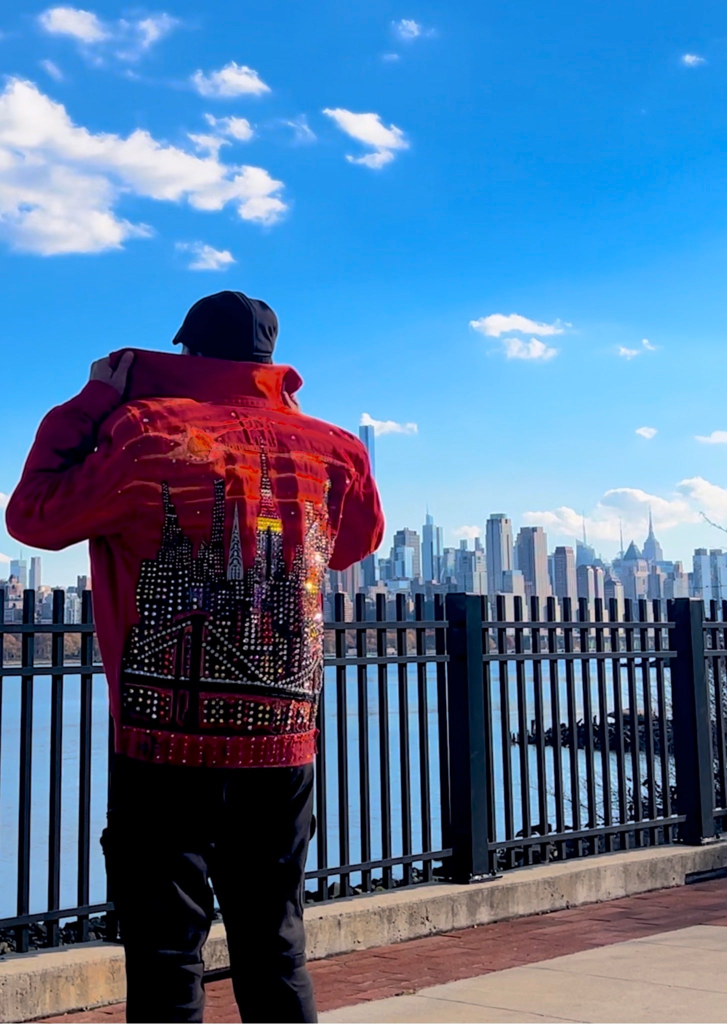 Person wearing a red jacket with a Loria NYC Art, standing on a bridge overlooking a city skyline.