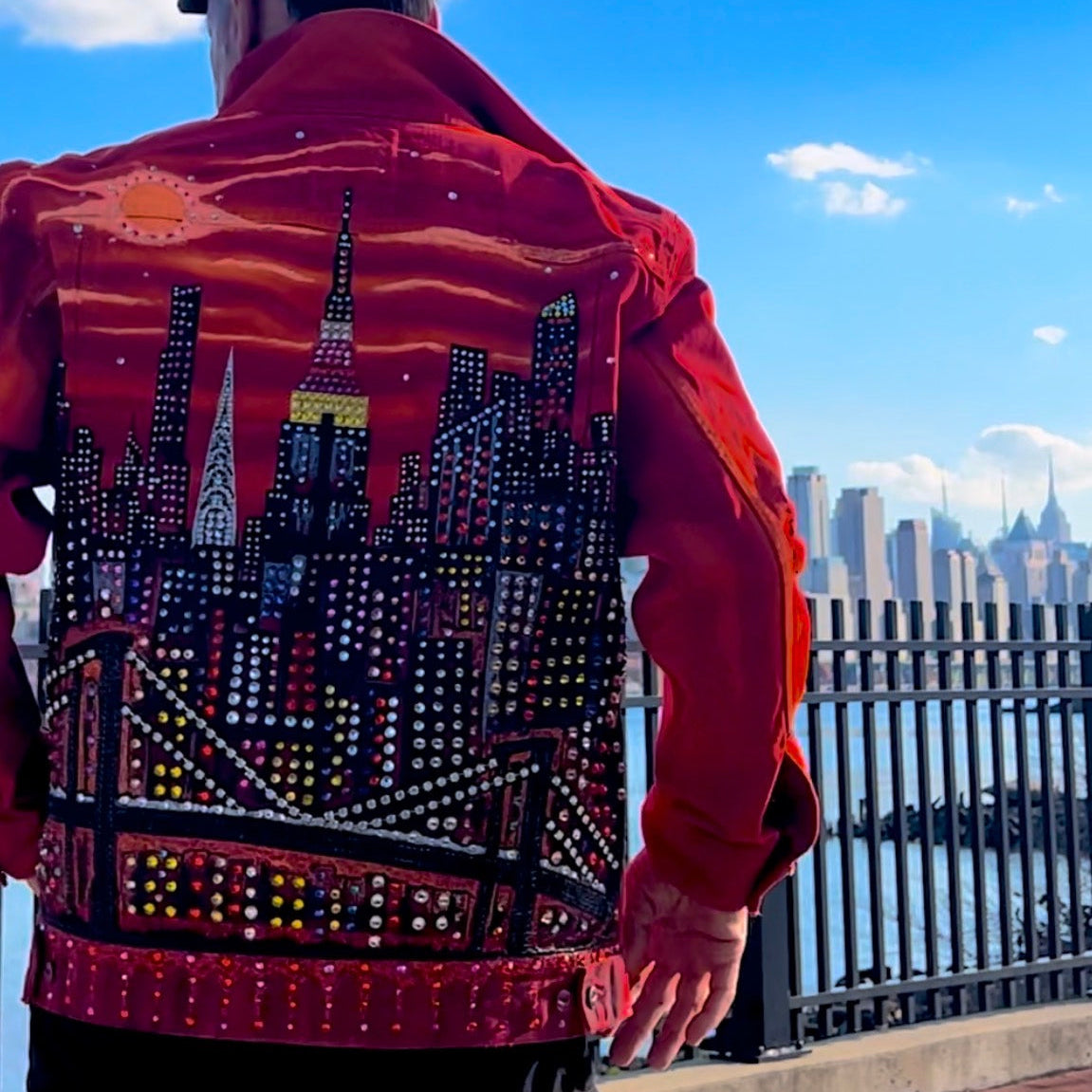 Person wearing a  jacket with a Loria NYC Art cityscape design, standing against a blue sky with clouds.