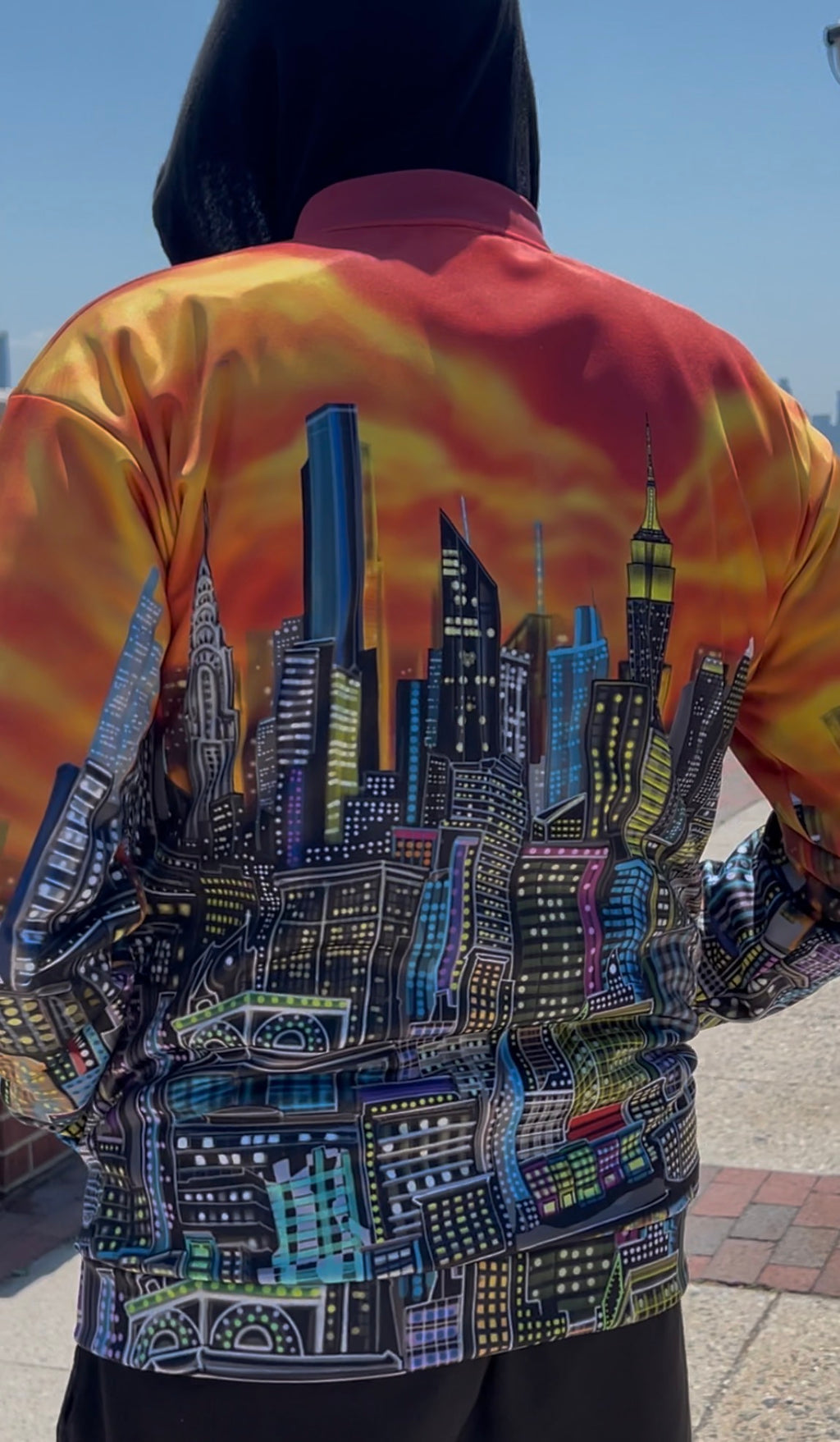 A person viewed from behind wearing a Loria NYC Art bomber-style jacket with a vibrant orange and yellow tie-dye flame pattern on the upper back and a detailed multicolor city skyline graphic printed across the lower back.