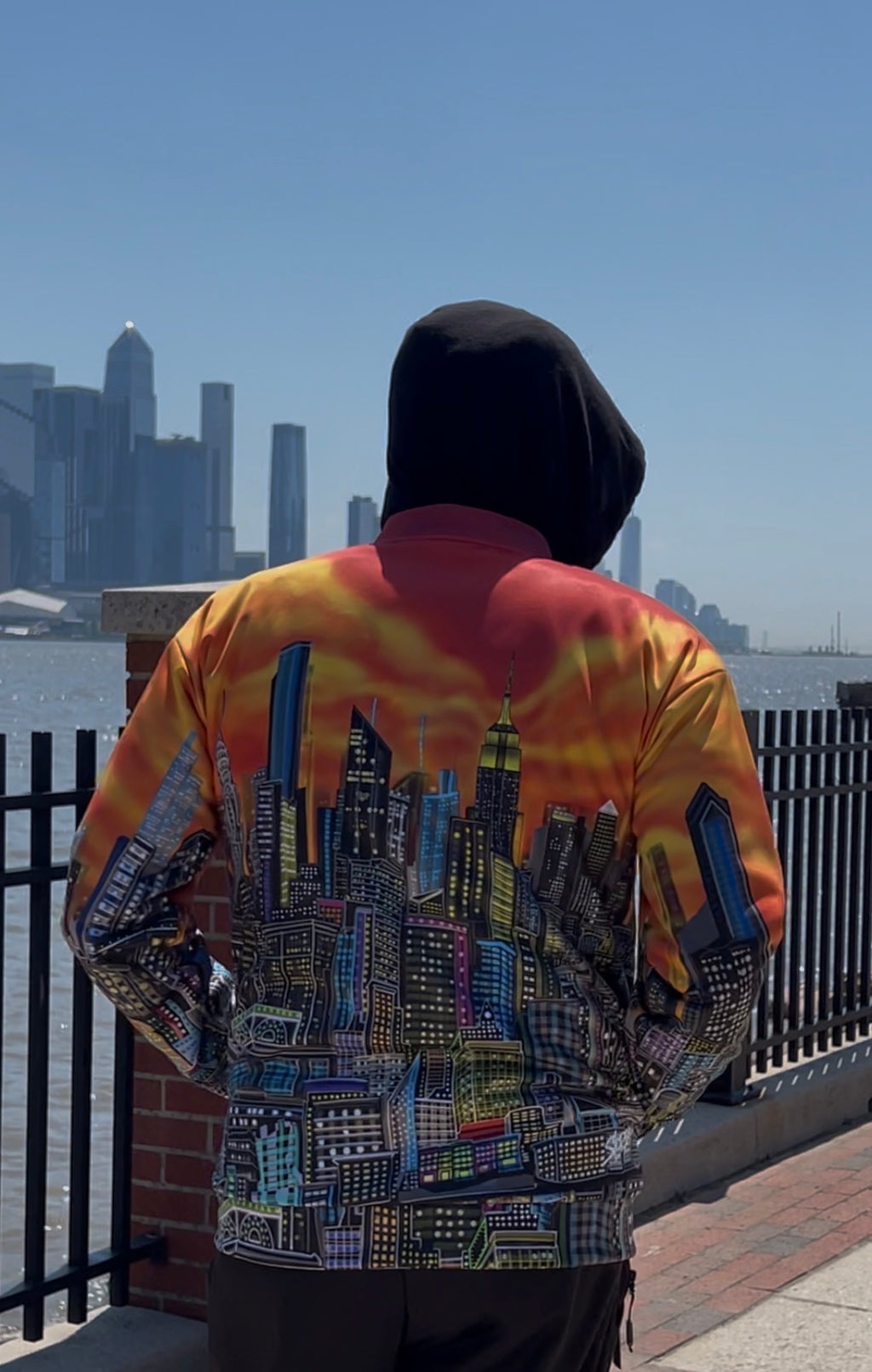 A person viewed from behind wearing a hooded jacket with a vibrant orange, yellow, and red sky print on the upper back and sleeves, and a detailed multicolor city skyline Loria NYC Art graphic across the lower back. The person is standing outdoors on a waterfront with a city skyline in the background under a clear blue sky.