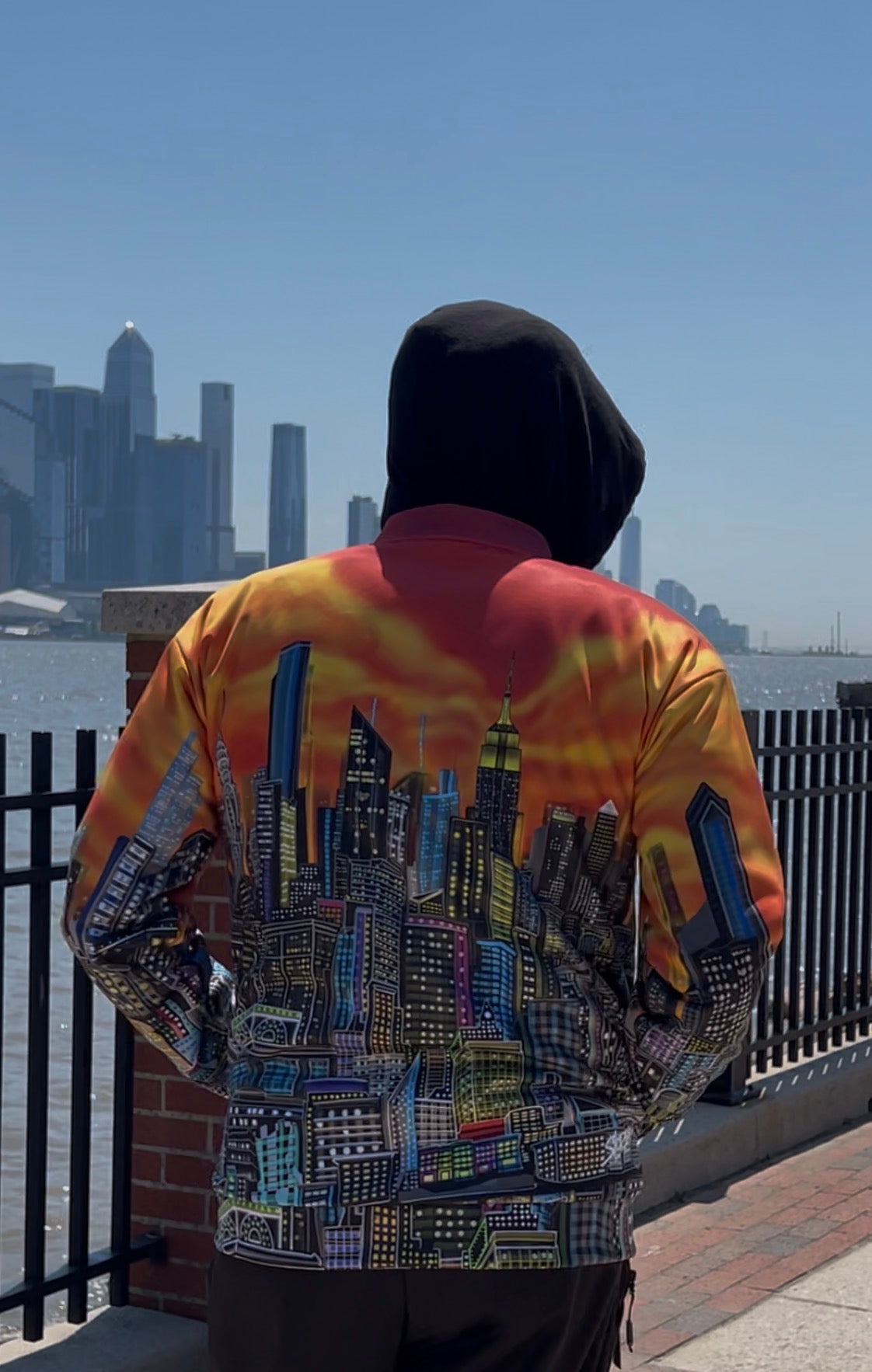 A person viewed from behind wearing a hooded jacket with a vibrant orange, yellow, and red sky print on the upper back and sleeves, and a detailed multicolor city skyline Loria NYC Art graphic across the lower back. The person is standing outdoors on a waterfront with a city skyline in the background under a clear blue sky.
