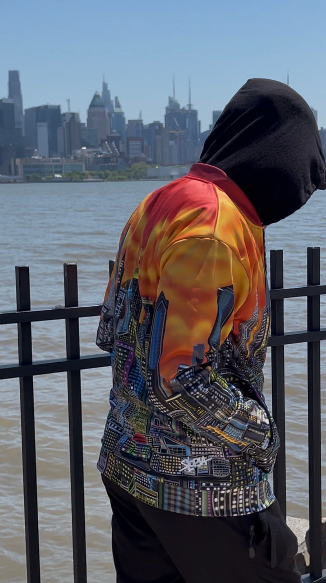 A person wearing a hooded jacket with a vibrant orange, yellow, and red sunset flame pattern on the upper back and a multicolor cityscape Loria NYC Art graphic on the lower back, standing by a waterfront with a city skyline in the background.