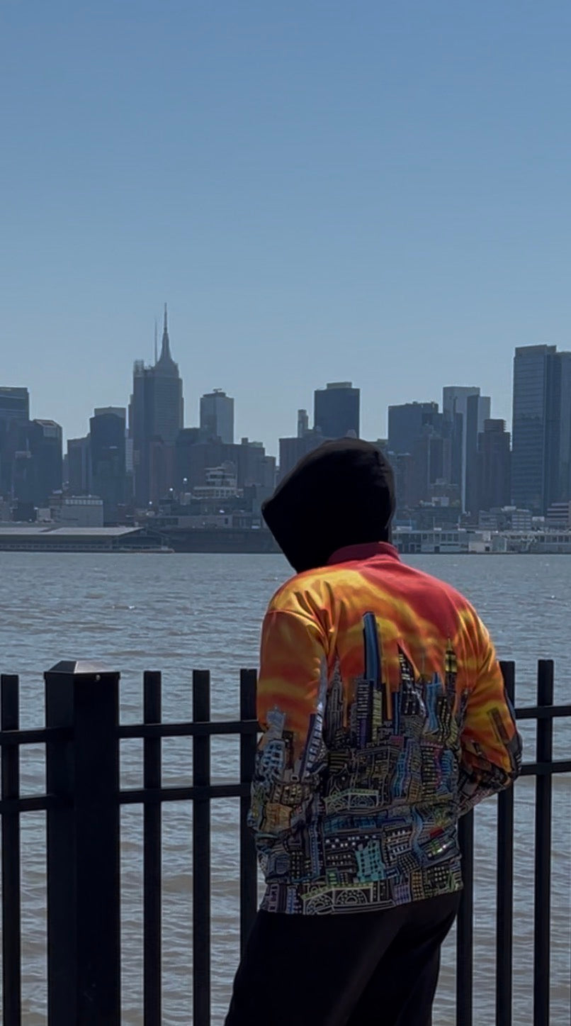 A person wearing a hooded sweatshirt and a vibrant orange, yellow, and red tie-dye graphic Loria NYC Art jacket stands with their back to the camera, looking out over a body of water toward a city skyline under a clear blue sky.