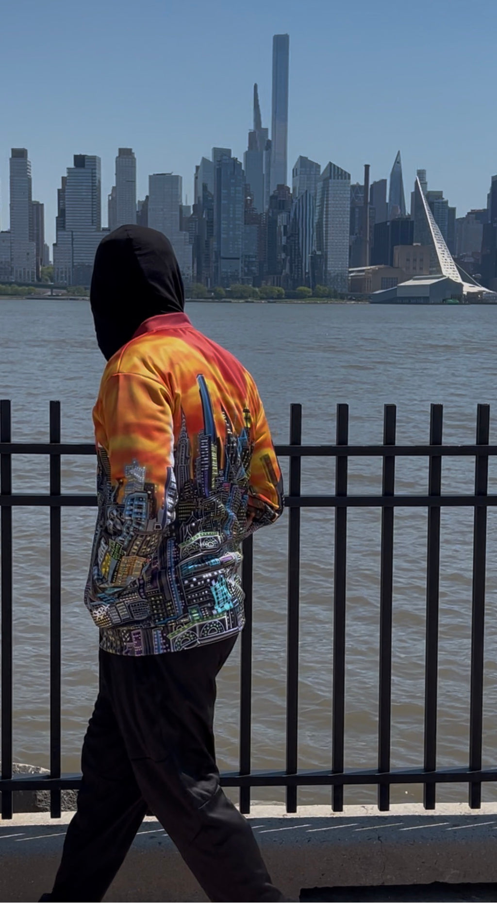 A person wearing a colorful Loria NYC Art graphic jacket and black pants walks along a waterfront railing with a city skyline in the background.