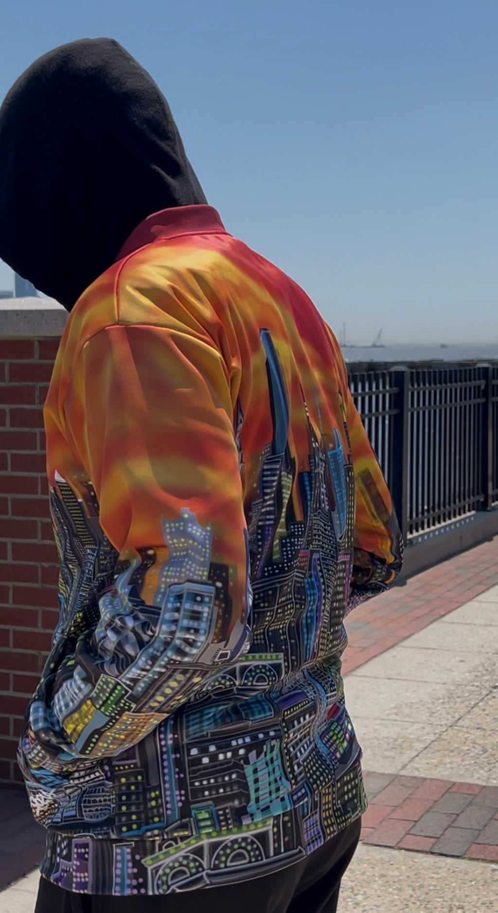 A person outdoors wearing a Loria NYC Art jacket with a fiery orange, yellow, and red gradient upper back and shoulders, transitioning into a detailed multicolor cityscape print on the lower back and sleeves.