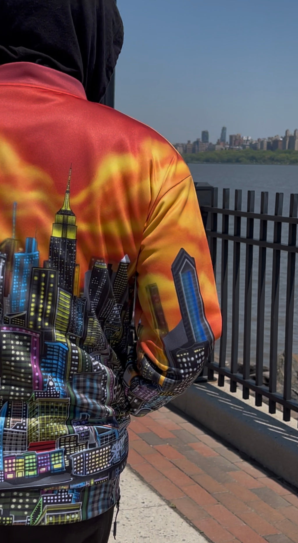 A person standing outdoors by a waterfront railing, wearing a colorful Loria NYC Art jacket with a fiery orange and yellow back design and a multicolored city skyline print on the front and sleeves.