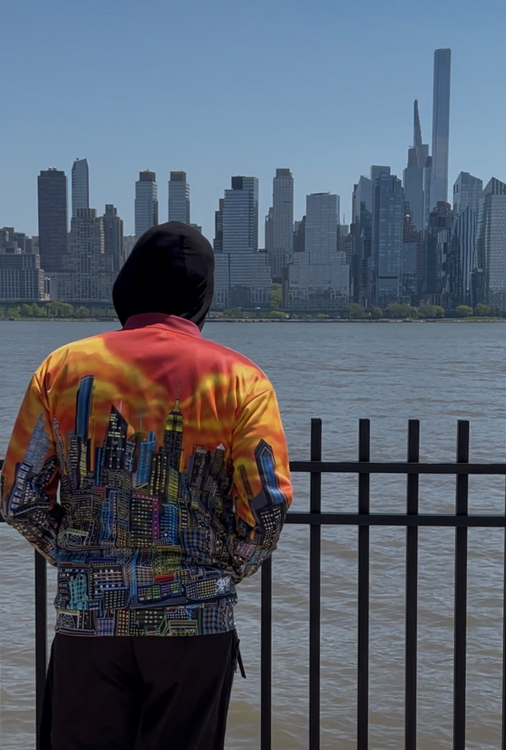 A person viewed from behind wearing a hooded jacket with a Loria NYC Art colorful city skyline print, standing by a waterfront fence with a city skyline in the background.