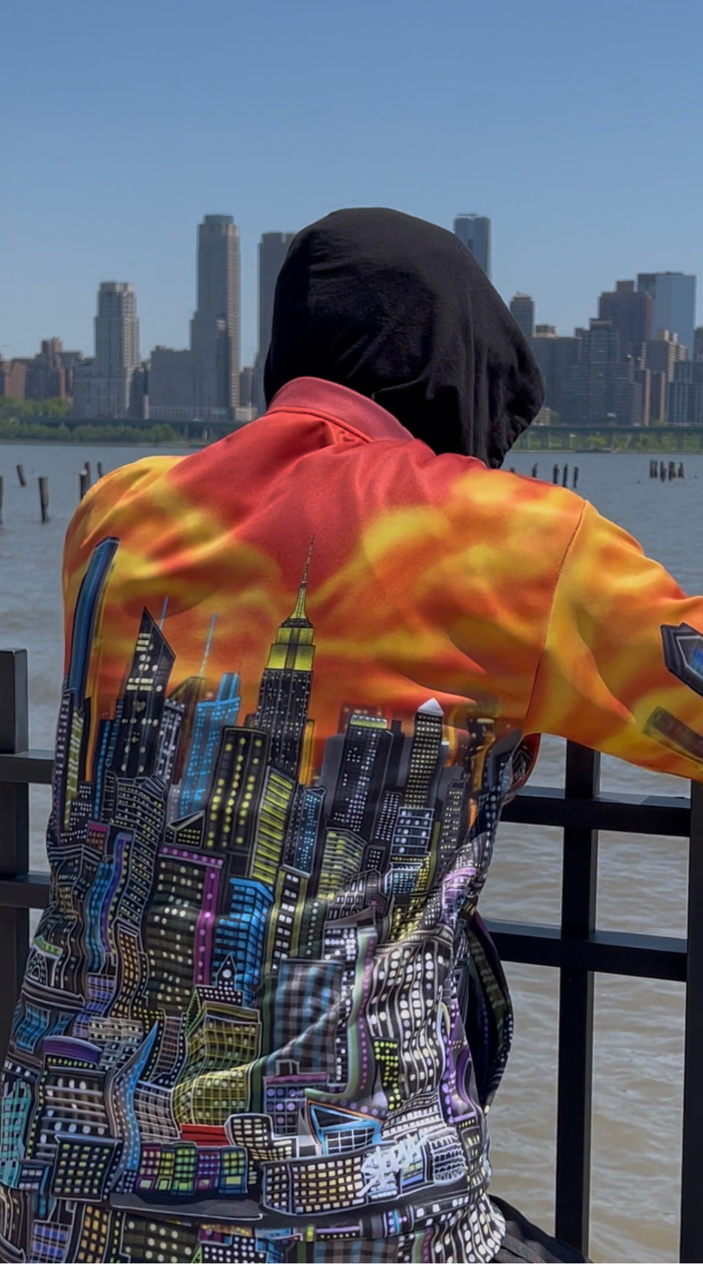 A person viewed from behind wearing a Loria NYC Art jacket with a vibrant orange and yellow flame pattern on the upper back and a multicolor city skyline graphic on the lower back, standing by a waterfront with a city skyline in the background.