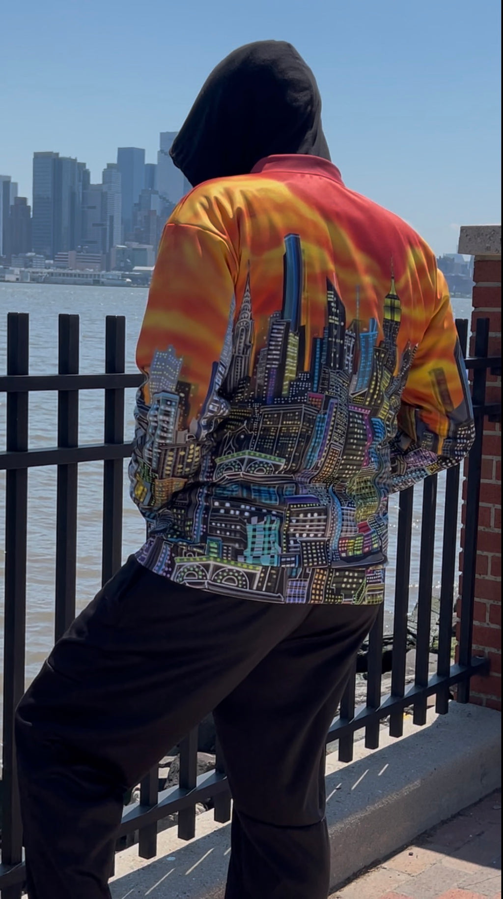 A person wearing a Loria NYC Art jacket with a vibrant orange and yellow sunset gradient on the upper back and a detailed multicolor city skyline print on the lower back, standing by a waterfront railing.