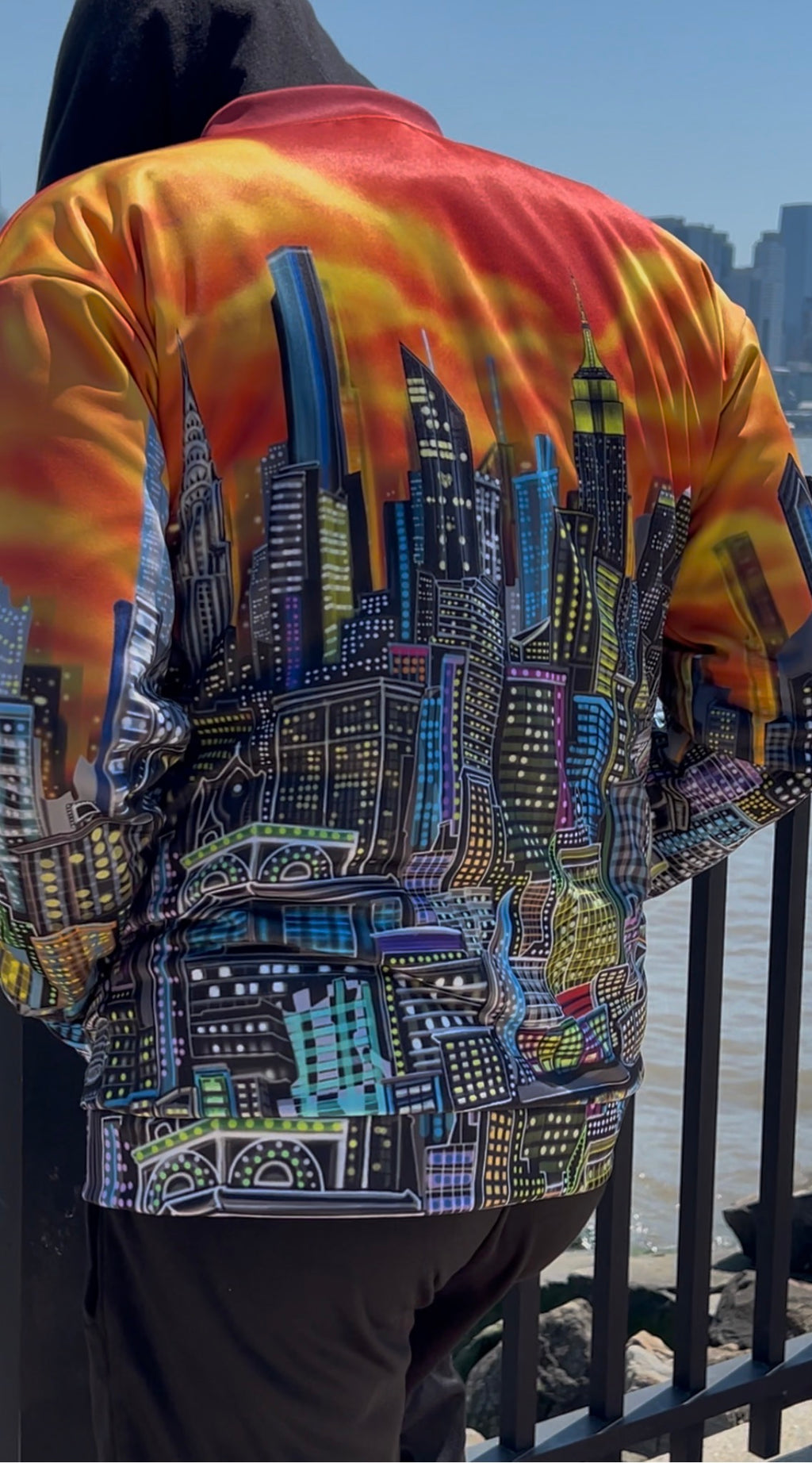 A person viewed from behind wearing a vibrant Loria NYC Art jacket with a detailed city skyline graphic across the back, featuring a fiery orange and yellow gradient sky. The person is standing outdoors near a railing.