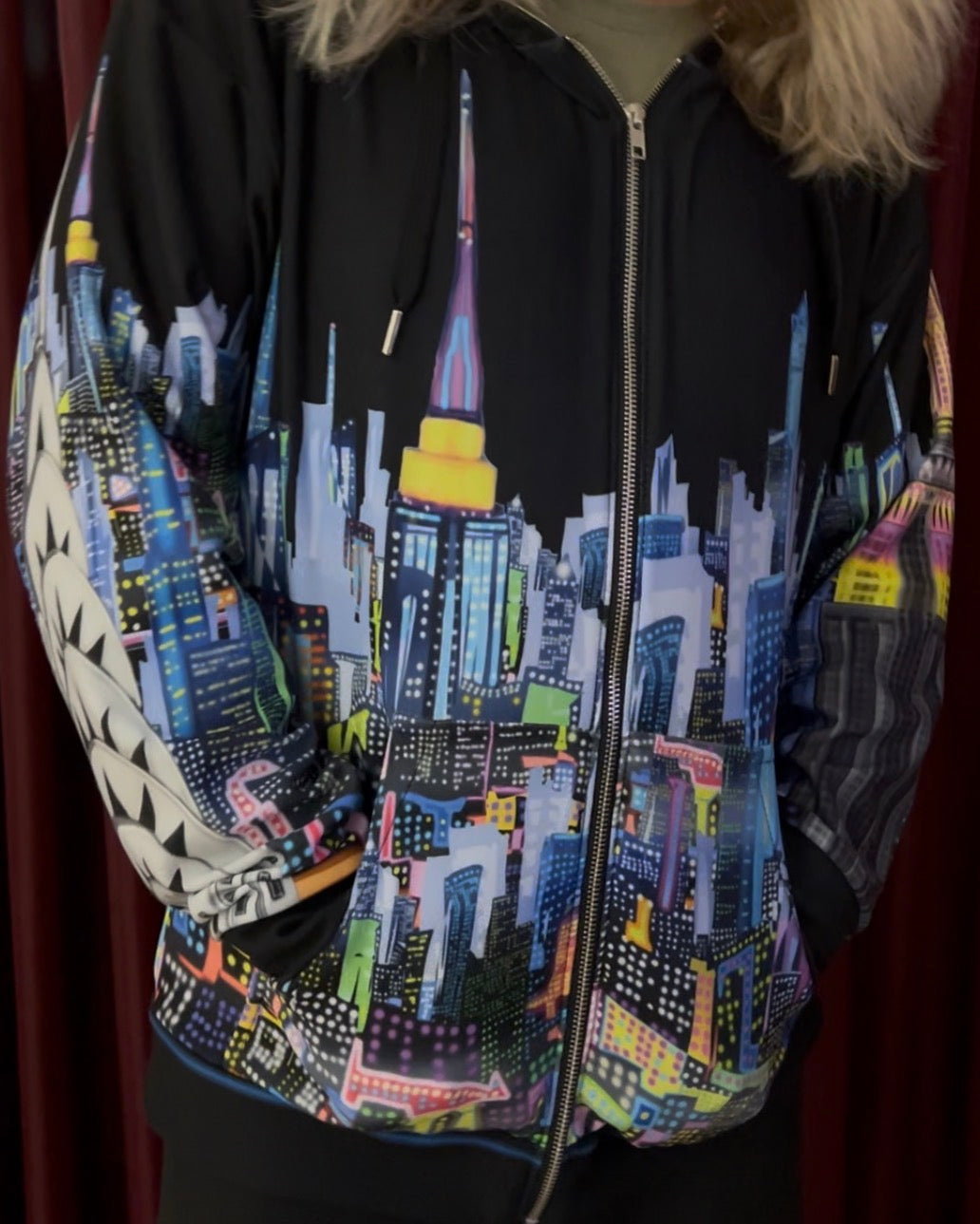 A person wearing a black zip-up hoodie with a colorful cityscape Loria NYC Art across the front and sleeves, featuring a stylized skyline with skyscrapers in blue, purple, pink, yellow, and white. The hoodie has a drawstring hood, front zipper, and ribbed cuffs and hem, shown against a dark red curtain background.