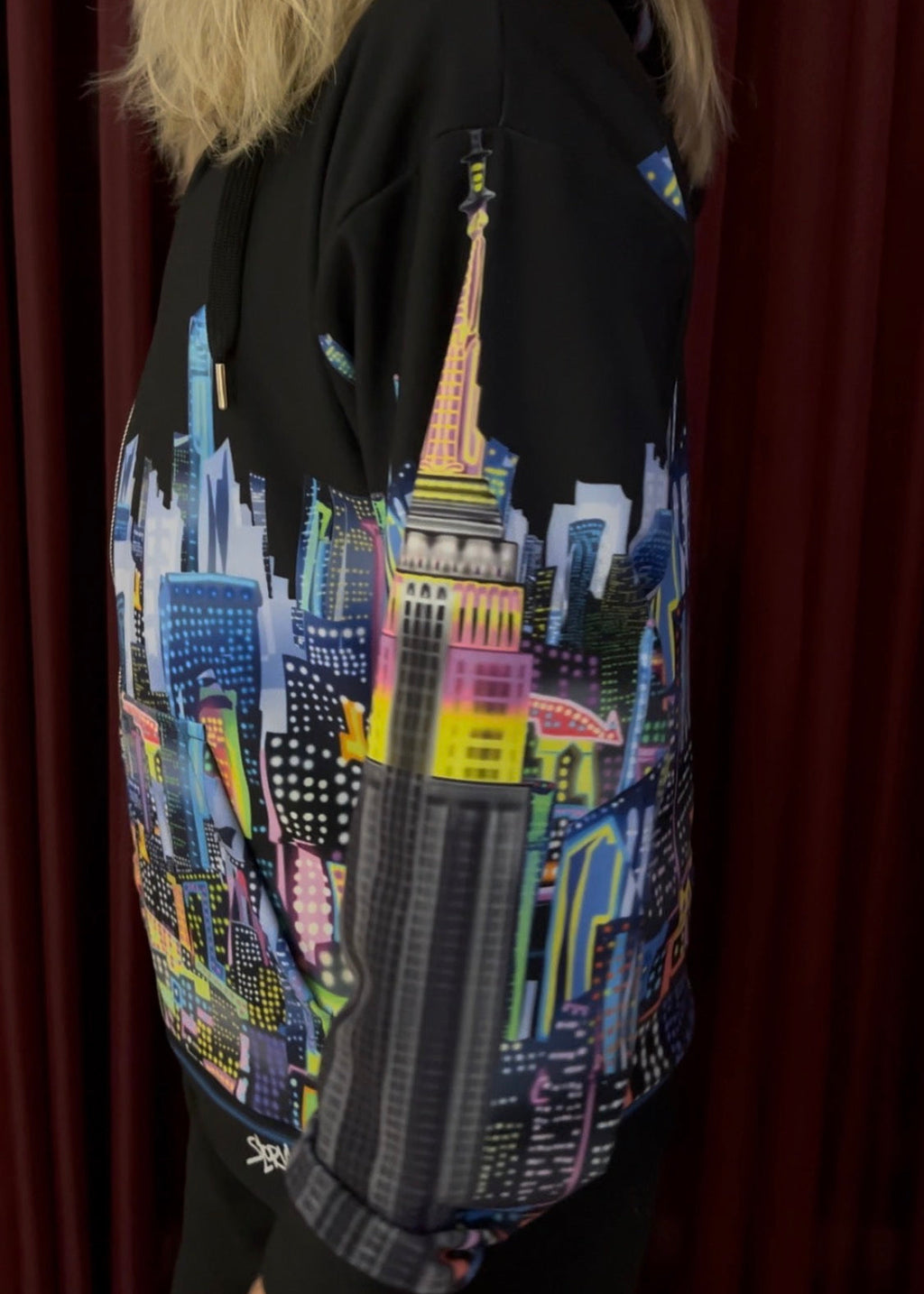 A person wearing a black hoodie with a vibrant, multicolored Loria NYC Art graphic print of a city skyline on the back, standing in front of dark red curtains.
