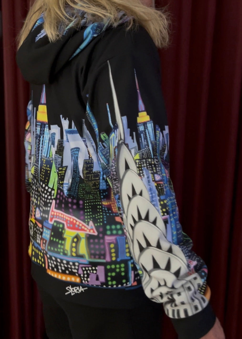 A person wearing a black hoodie with a colorful city skyline Loria NYC Art graphic across the back and a shark-face pattern on the sleeve, photographed against a dark red curtain background.