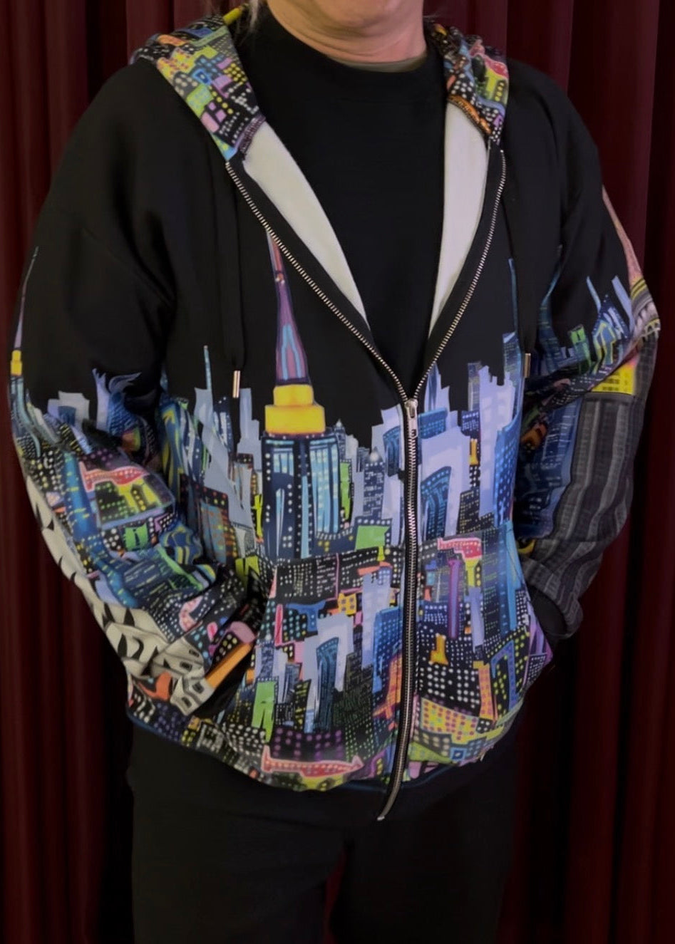 A person wearing a black zip-up hoodie with a colorful Loria NYC Art skyline print on the front, sleeves, and hood, paired with a black t-shirt and black pants, standing in front of a dark red curtain.