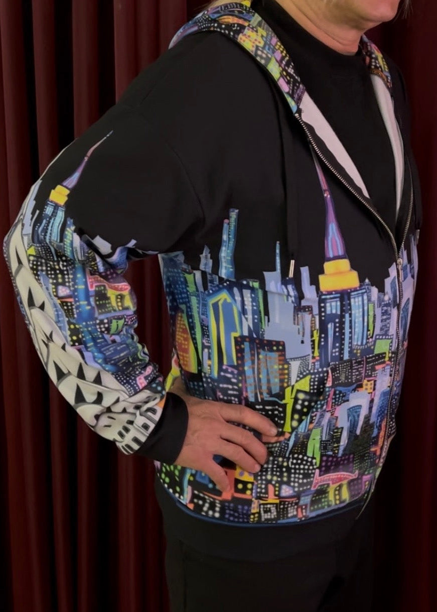 A person wearing a black zip-up hoodie with a colorful city skyline Loria NYC Art print on the front and sleeves, paired with a black t-shirt, posed against a dark red curtain background.