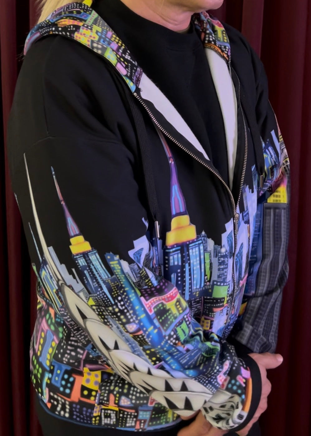 A person wearing a black zip-up hoodie with a colorful cityscape Loria NYC Art print on the hood, shoulders, and sleeves, layered over a black shirt. The background is a dark red curtain.
