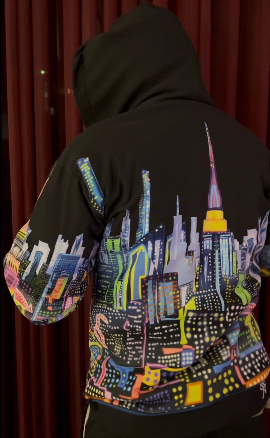 A person wearing a black hoodie with a colorful, vibrant city skyline Loria NYC Art graphic printed across the back and sleeves, photographed against a dark red curtain background.