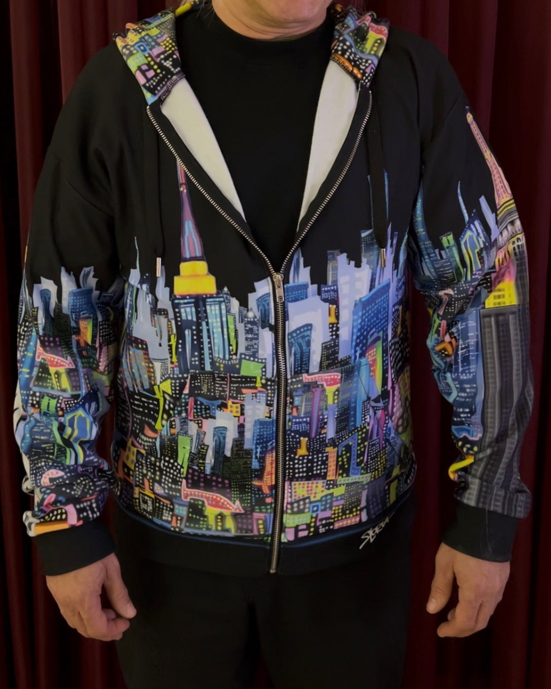 A man wearing a black zip-up hoodie with a vibrant, multicolored New York City skyline Loria NYC Art graphic printed across the front and sleeves, paired with a black t-shirt and black pants, standing in front of a dark red curtain.