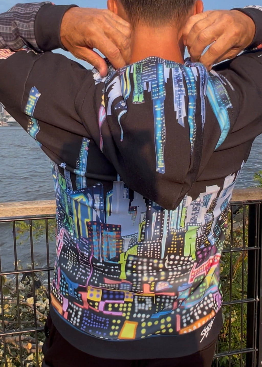 A man viewed from behind wearing a black Loria NYC Art hoodie with a vibrant multicolor neon cityscape graphic print on the back, standing outdoors near a railing with water in the background.