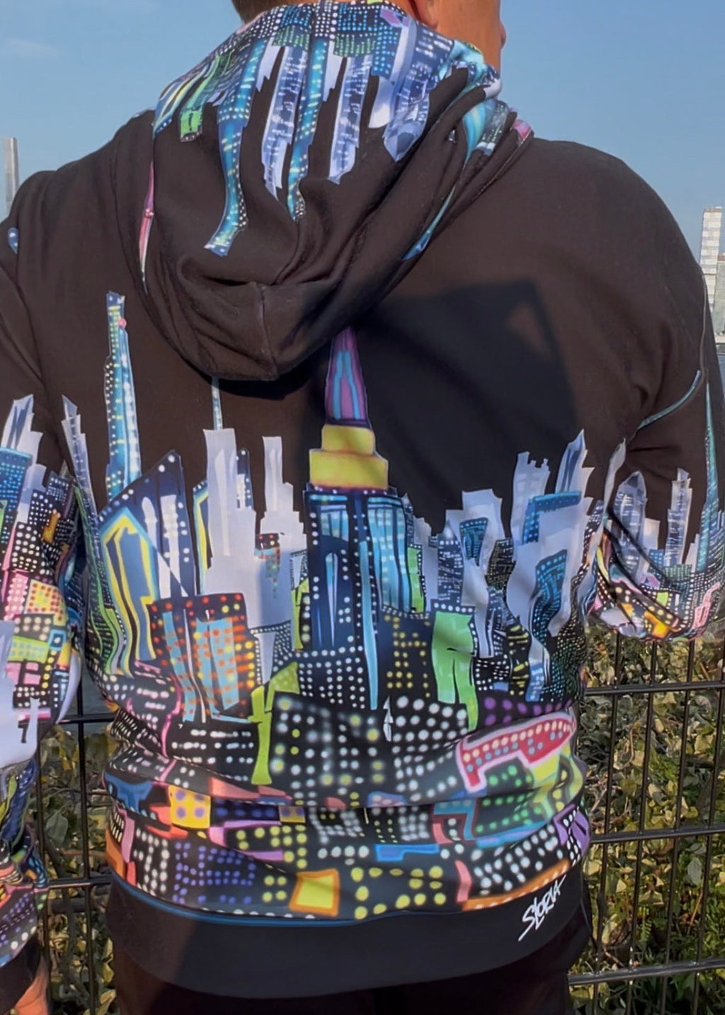 A man viewed from behind wearing a black Loria NYC Art hoodie with a vibrant, multicolored cityscape graphic printed across the back and sleeves, paired with black pants.