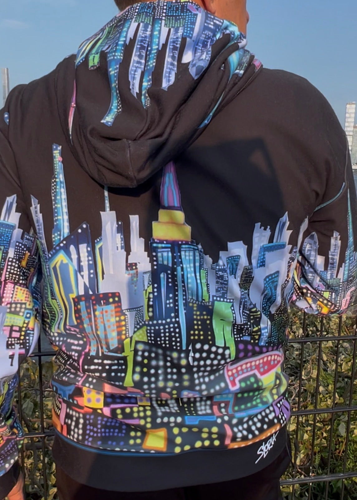 A man viewed from behind wearing a black Loria NYC Art hoodie with a vibrant, multicolored cityscape graphic printed across the back and sleeves, paired with black pants.