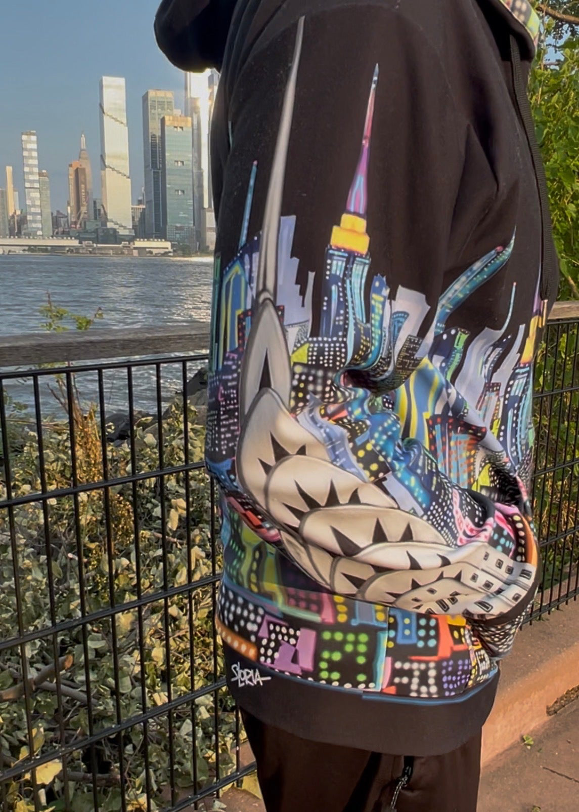 A person wearing a black hoodie with a highly detailed, colorful Loria NYC Art graphic print of the New York City skyline, standing outdoors near a railing with a city skyline and river in the background.