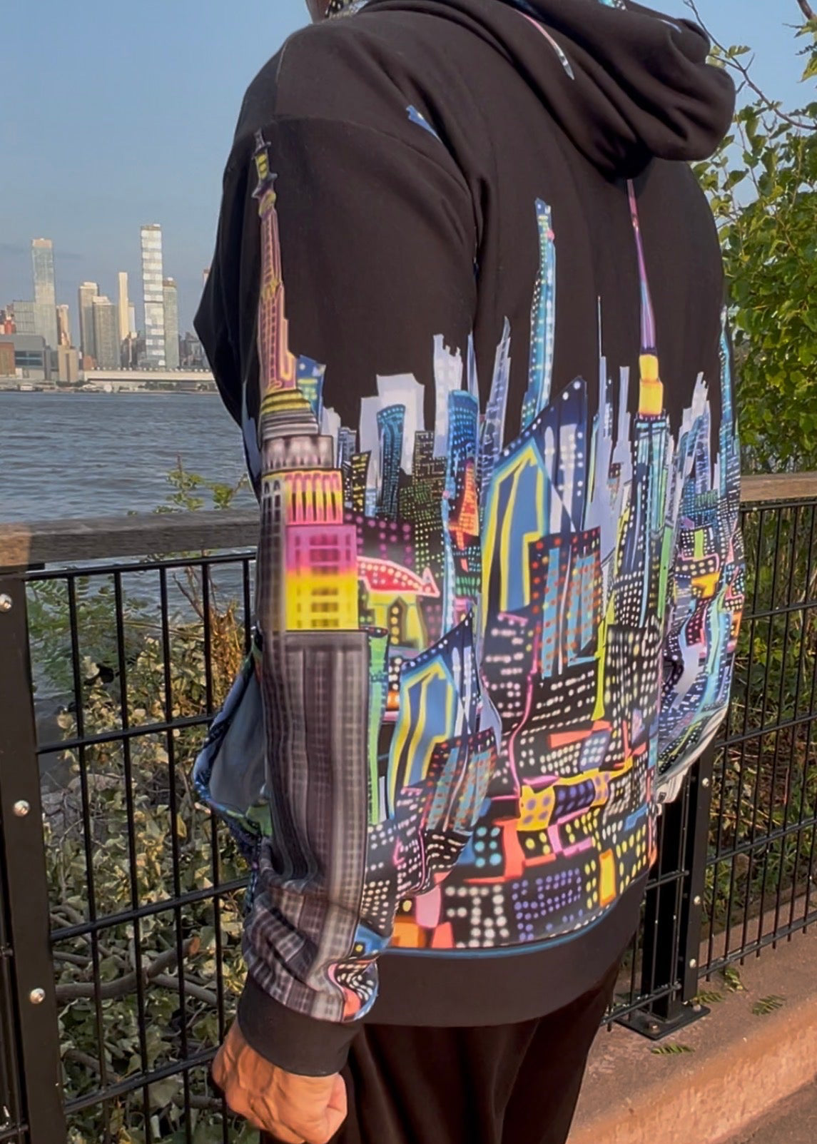 A person wearing a black Loria NYC Art hoodie with a large, colorful graphic print of a city skyline on the back, standing outdoors next to a railing with water and buildings in the background.