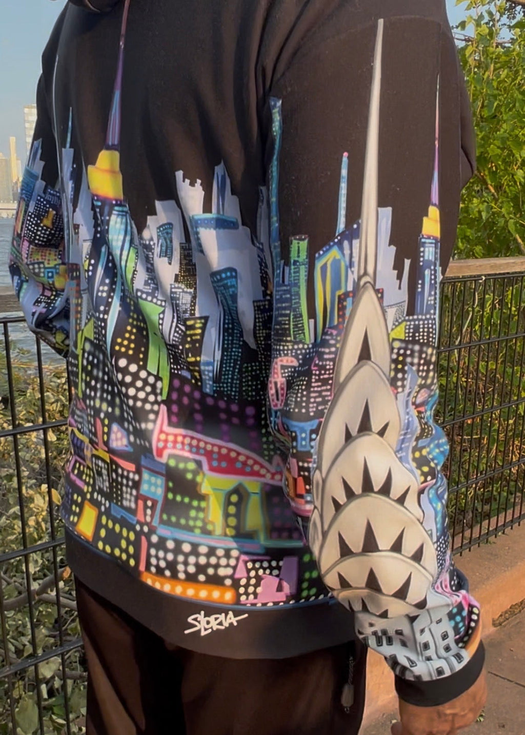 A person wearing a black Loria NYC Art hoodie with a colorful city skyline graphic across the back and sleeves, photographed outdoors near a railing.