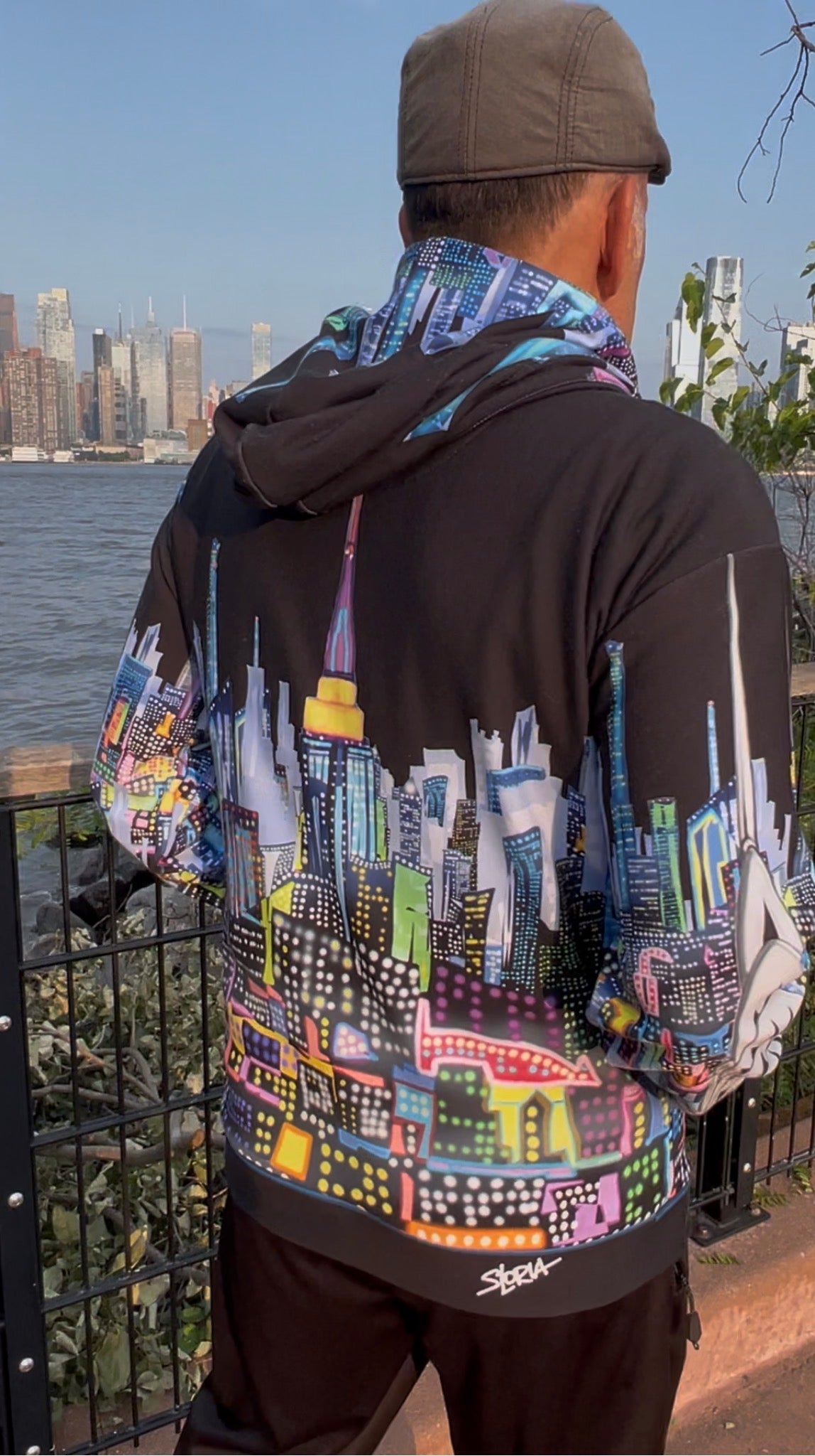 A man viewed from behind wearing a black hoodie with a vibrant, multicolored New York City skyline Loria NYC Art graphic across the back, standing outdoors near a waterfront railing.
