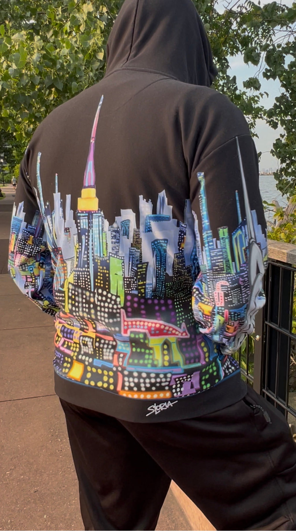 A person outdoors wearing a black hoodie with a large colorful city skyline Loria NYC Art graphic printed across the back and sleeves, paired with black pants.