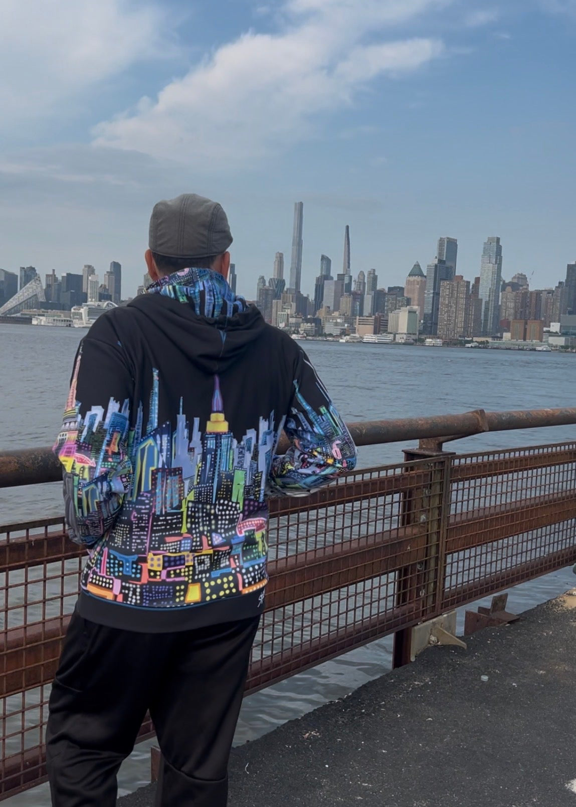 A man stands on a waterfront promenade wearing a black hoodie with a vibrant, Loria NYC Art skyline graphic on the back, paired with black pants and a gray cap, looking out over the water toward the Manhattan skyline.