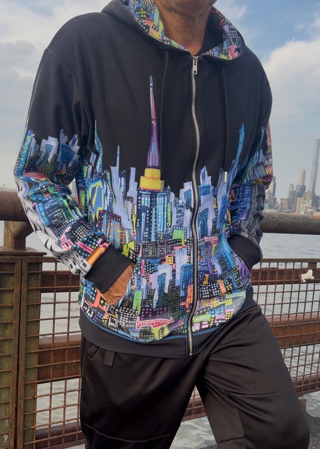 A person wearing a black zip-up Loria NYC Art hoodie with a colorful city skyline graphic across the front and sleeves, paired with black pants, standing outdoors near a railing and water.