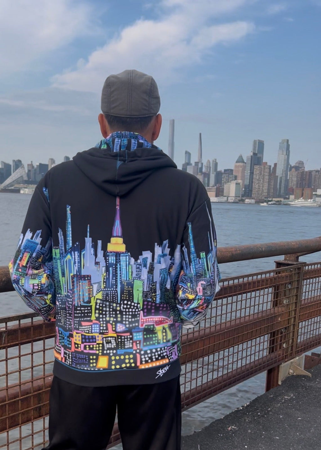 A man viewed from behind wearing a black hoodie with a vibrant, multicolored New York City skyline graphic on the back, standing on an outdoor pier with the city skyline and water in the background.