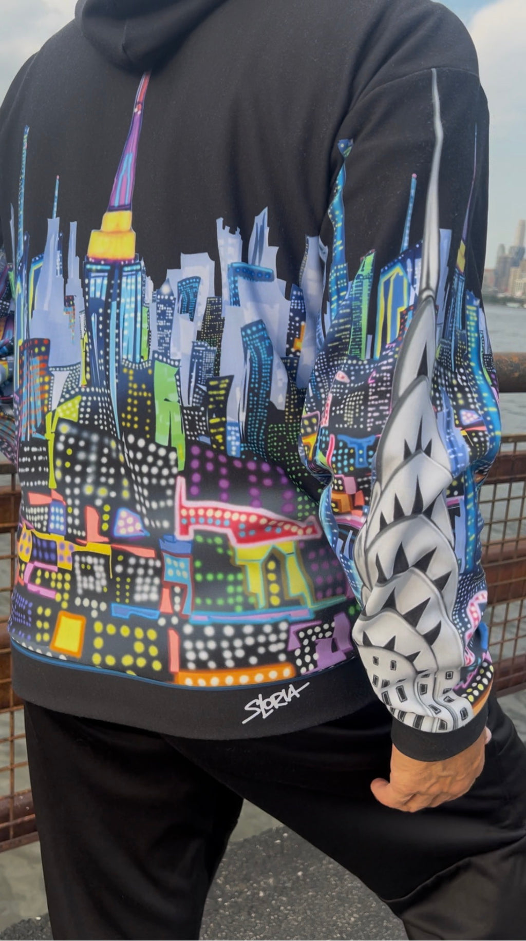 A person wearing a black Loria NYC Art hoodie with a colorful, stylized cityscape graphic across the back and sleeves, paired with black sweatpants. The hoodie features a vibrant multicolor print resembling a New York City skyline with neon lights, and a white geometric pattern on one sleeve.