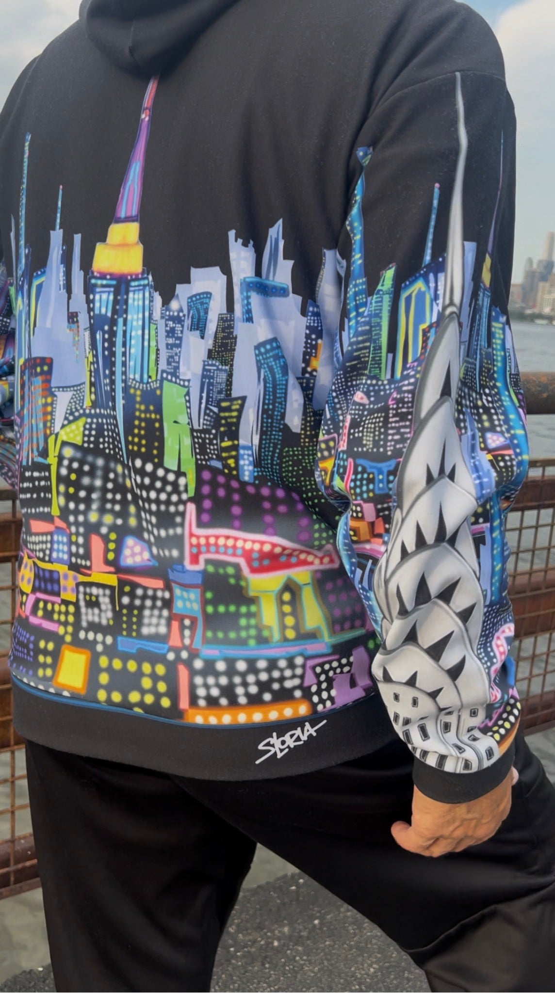 A person wearing a black Loria NYC Art hoodie with a colorful, stylized cityscape graphic across the back and sleeves, paired with black sweatpants. The hoodie features a vibrant multicolor print resembling a New York City skyline with neon lights, and a white geometric pattern on one sleeve.