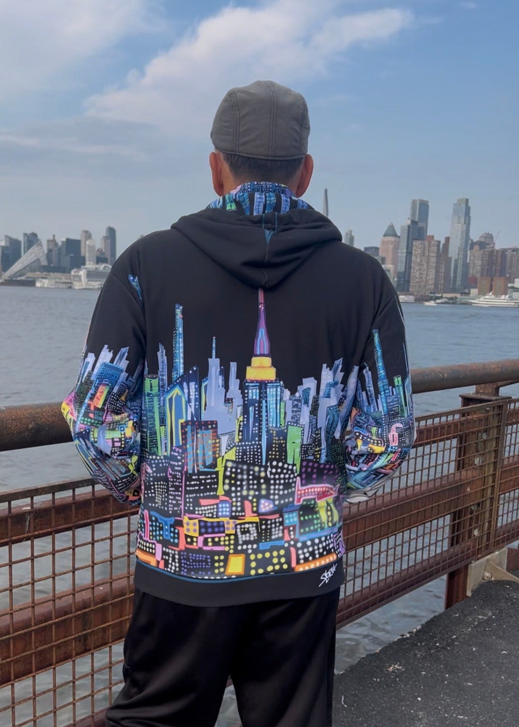 A man viewed from behind wearing a black Loria NYC Art hoodie with a vibrant, multicolored New York City skyline graphic across the back and sleeves, standing on a waterfront with a city skyline in the background.