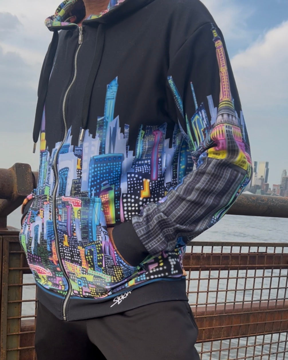 A person wearing a black zip-up Loria NYC Art hoodie with a colorful city skyline graphic across the front and multicolor plaid sleeves, paired with black pants, standing outdoors near a railing.