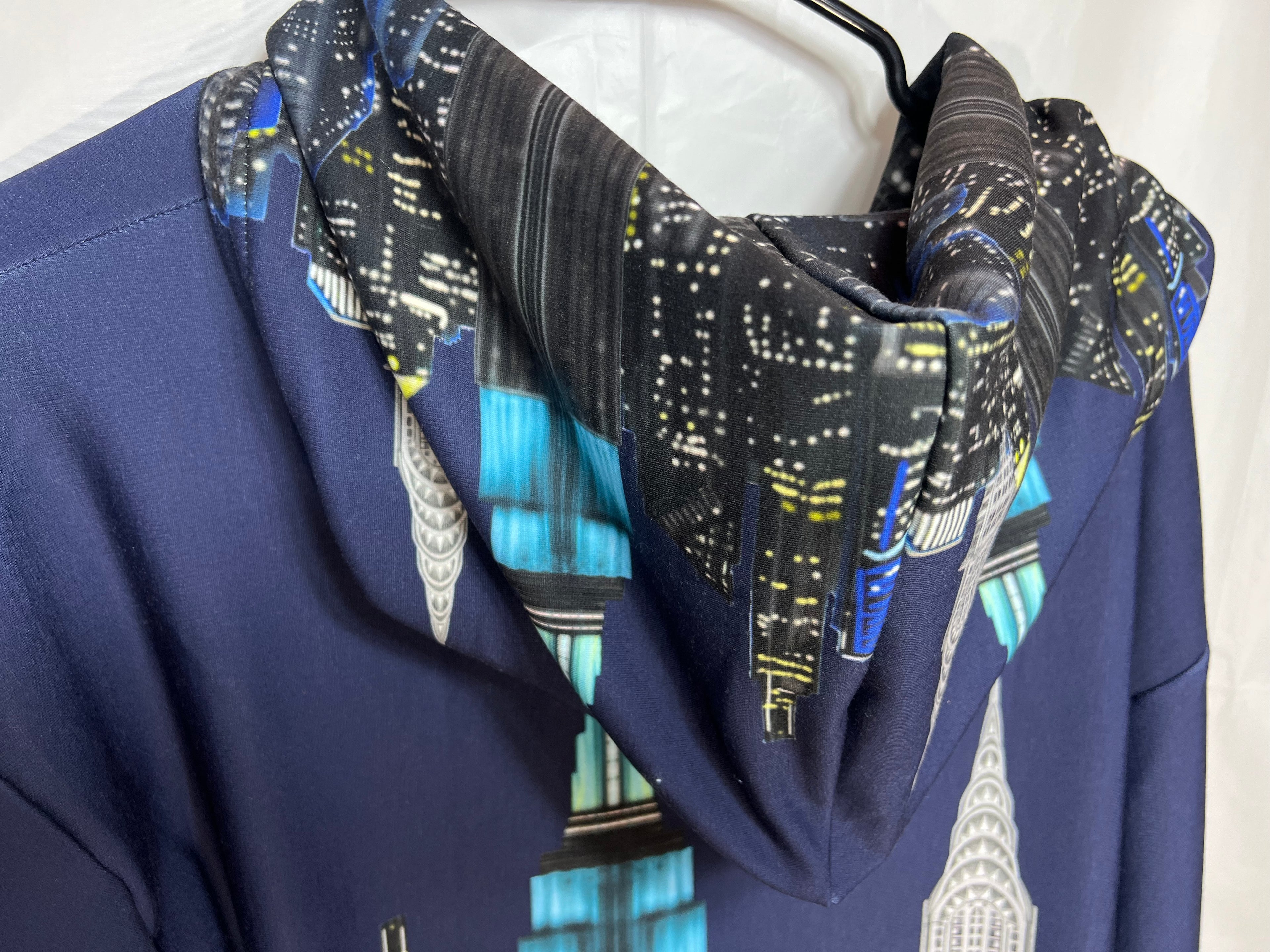 Navy blue garment with a Loria NYC Art cityscape hood draped over hoodie on a white background