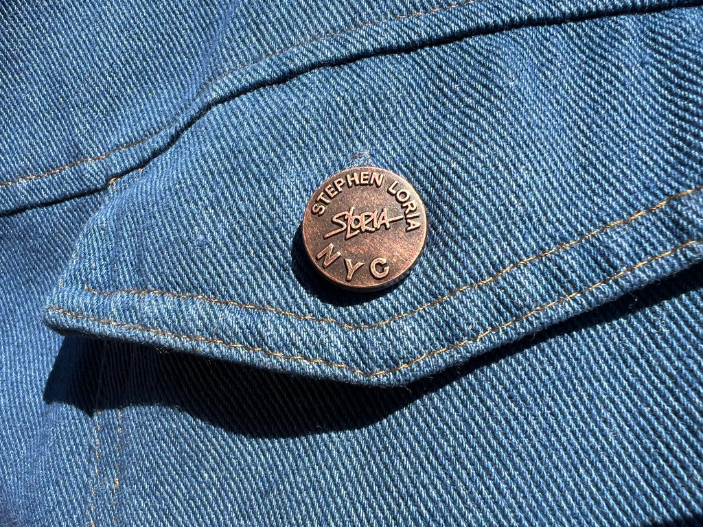Close-up of a blue denim jacket with a button featuring 'Stephen Loria' branding.