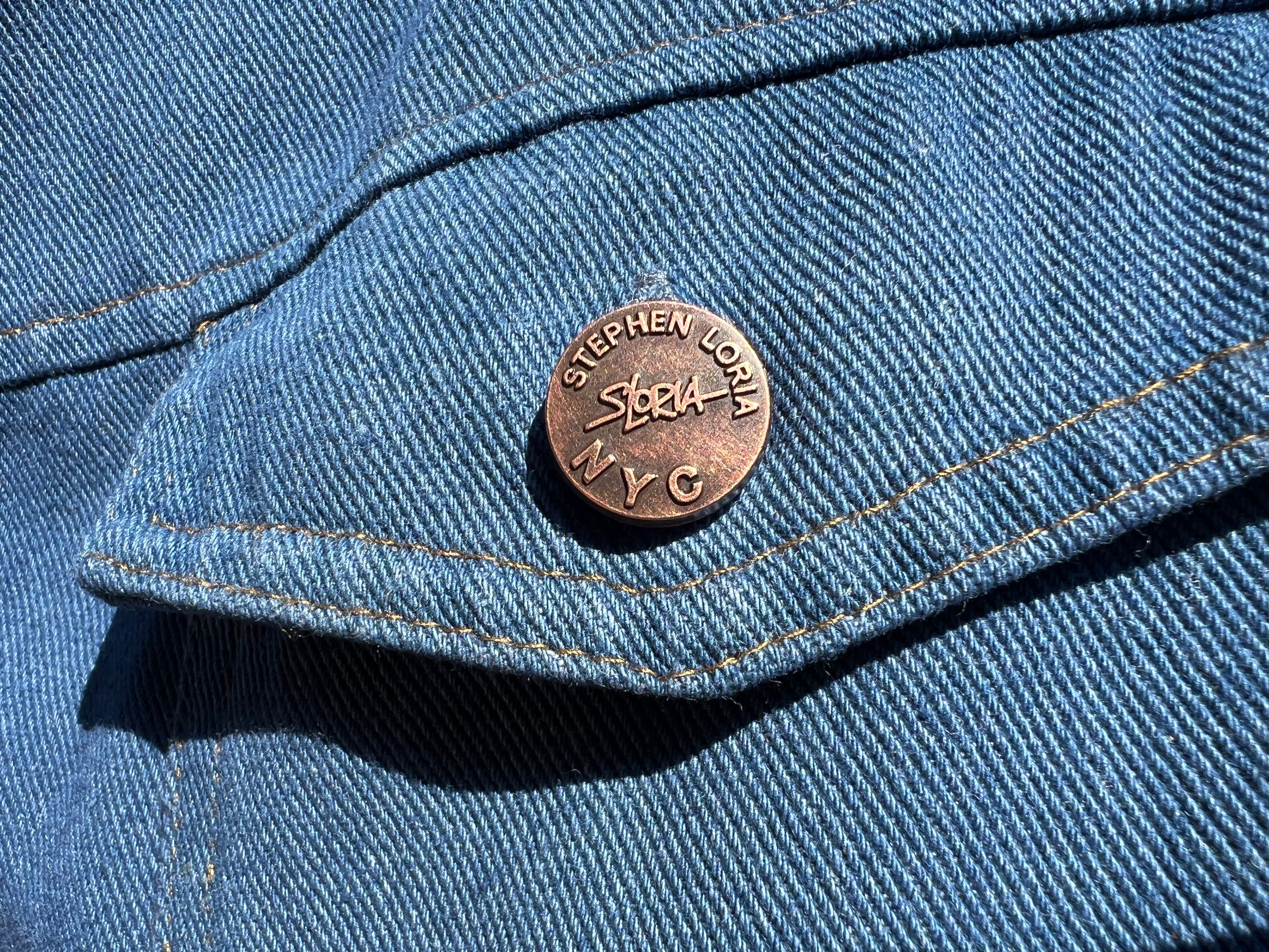 Close-up of a blue denim jacket with a button featuring 'Stephen Loria' branding.
