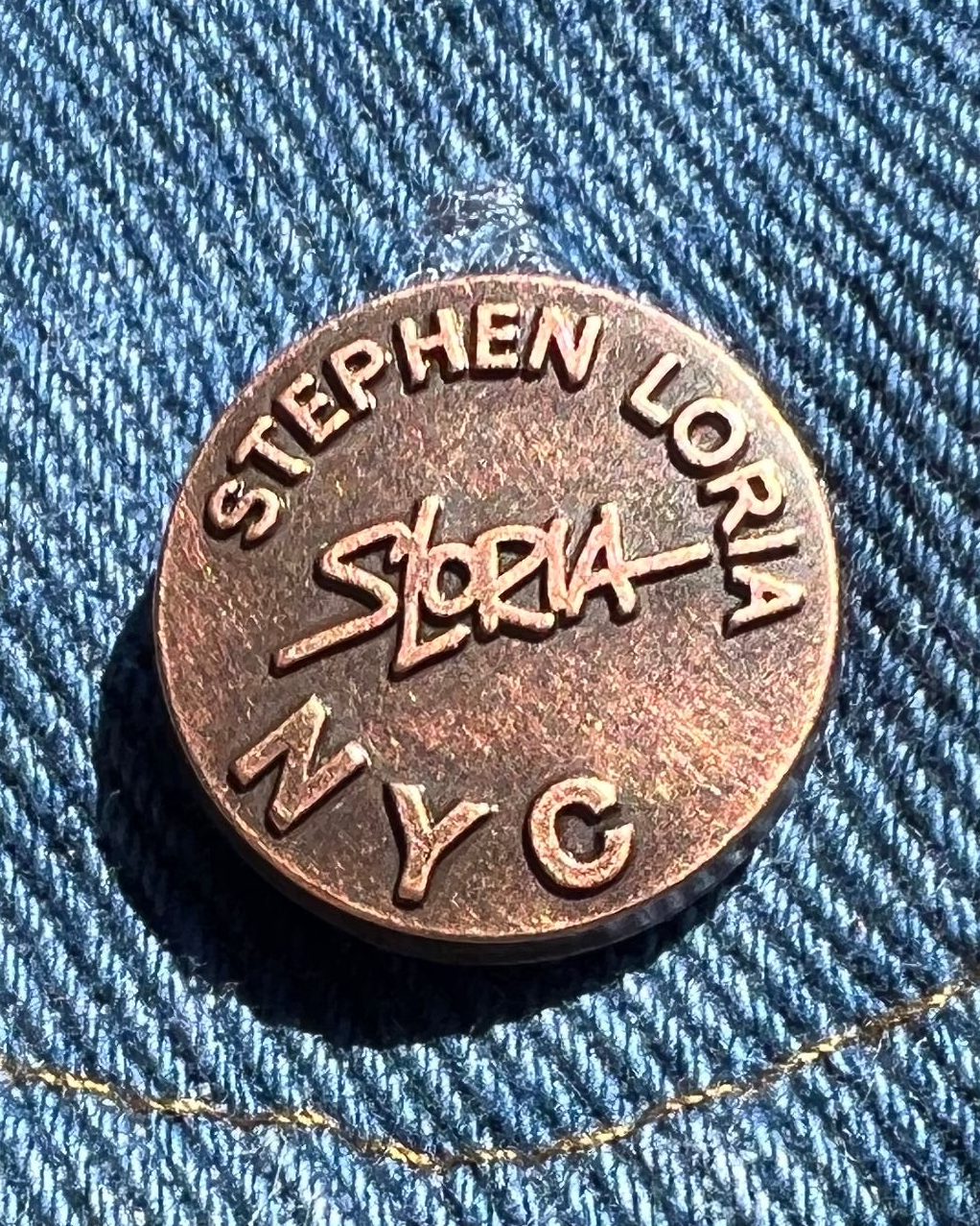 Round metal tag with 'Stephen Loria NYC' on a blue denim background